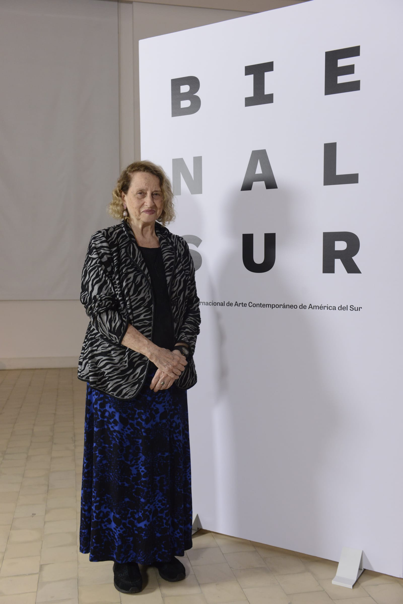 Anna Bella Geiger, una mujer mayor de pelo rubio rizado, con chaqueta de estampado cebra y falda azul, posa de pie junto a un panel blanco con el rótulo Bienal Sur.