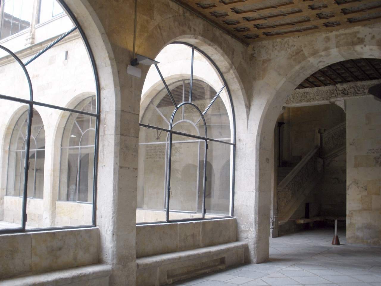 Galería de arcos de piedra con grandes ventanales de hierro que dan a un claustro, y al fondo una escalera de piedra tallada, en las Escuelas Mayores de la Universidad de Salamanca.