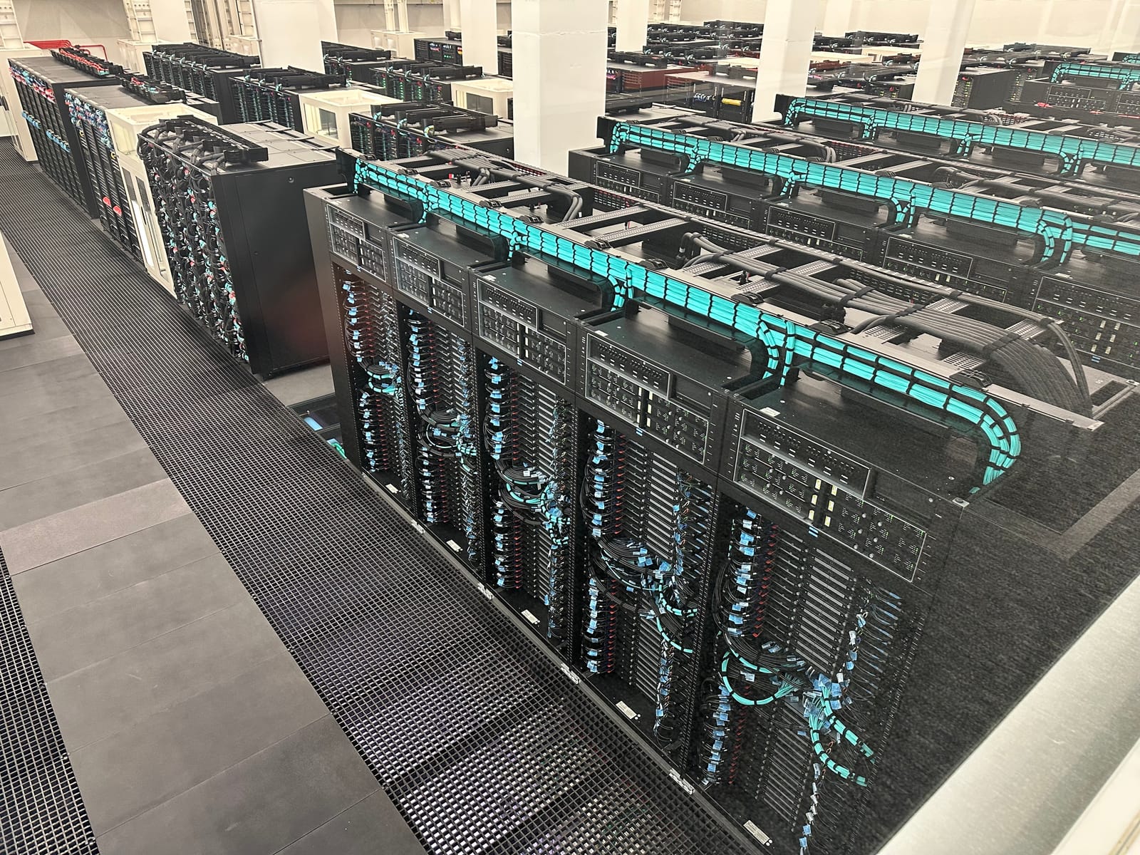 Filas de armarios negros del supercomputador MareNostrum 5, con mazos de cables azules y turquesa, vistas desde una pasarela de rejilla en la sala del Barcelona Supercomputing Center.