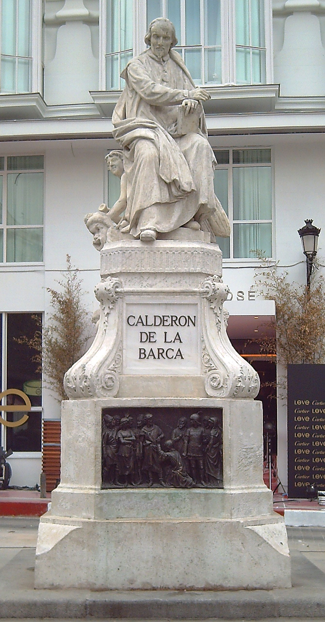 Estatua de piedra blanca de un hombre sentado con un libro, sobre un pedestal con la inscripción «Calderón de la Barca» y un relieve de bronce, frente a la fachada de un edificio.