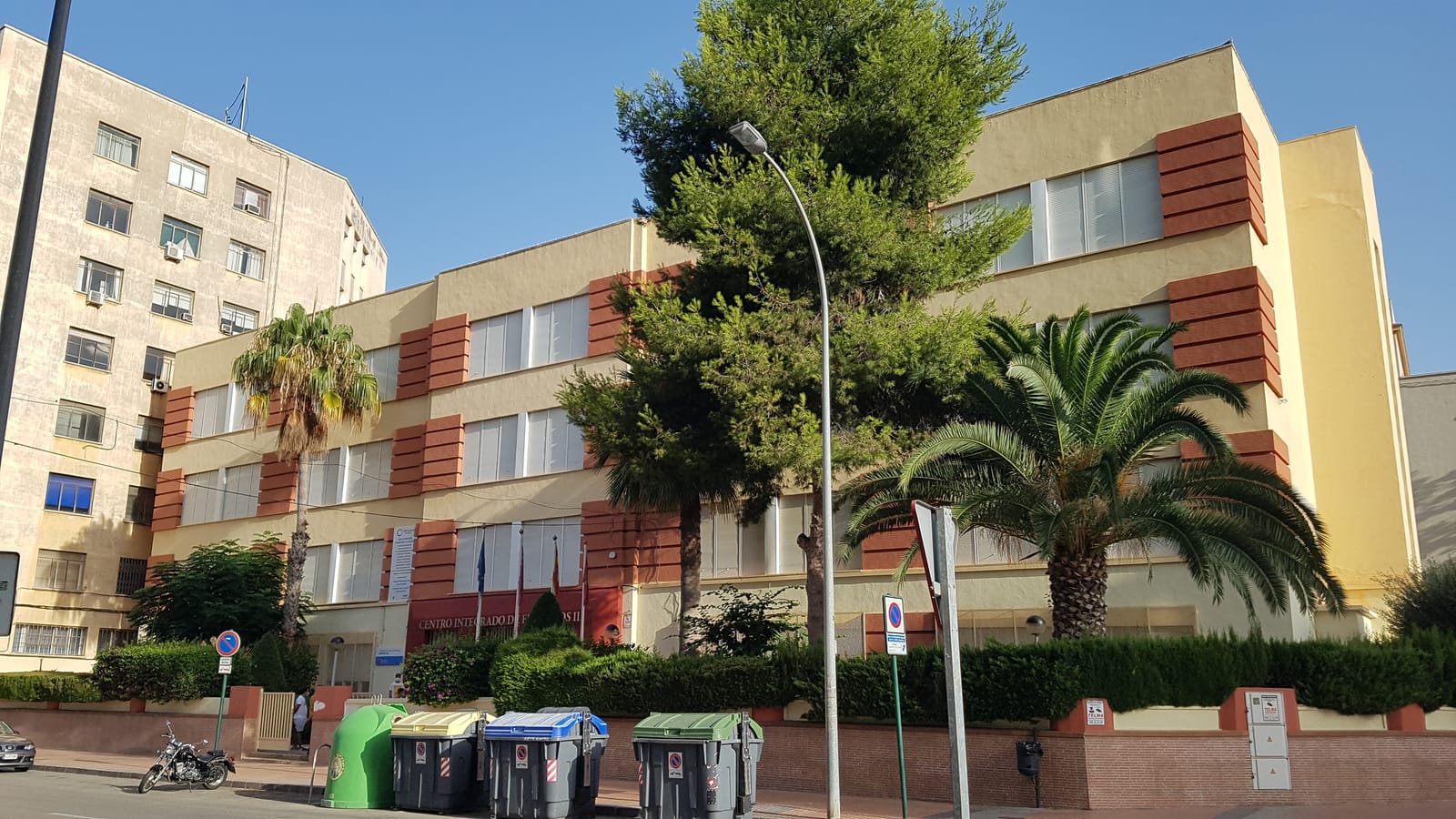 Fachada de tres plantas de un centro de formación profesional, con paños de ladrillo rojizo, palmeras y pinos en la acera y contenedores de reciclaje junto a la entrada.