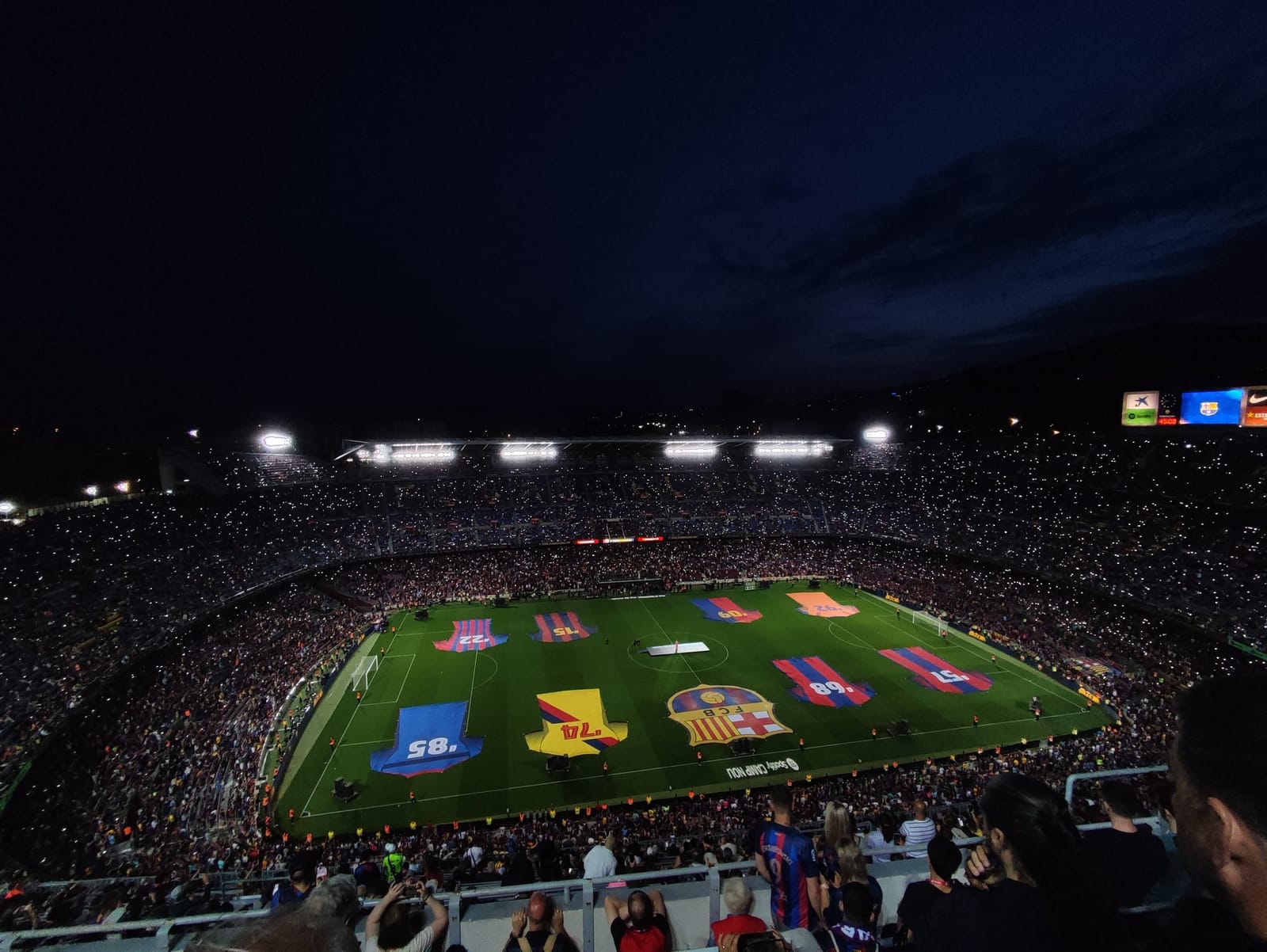 Vista nocturna del Camp Nou lleno de aficionados, con el césped decorado con camisetas gigantes del FC Barcelona.