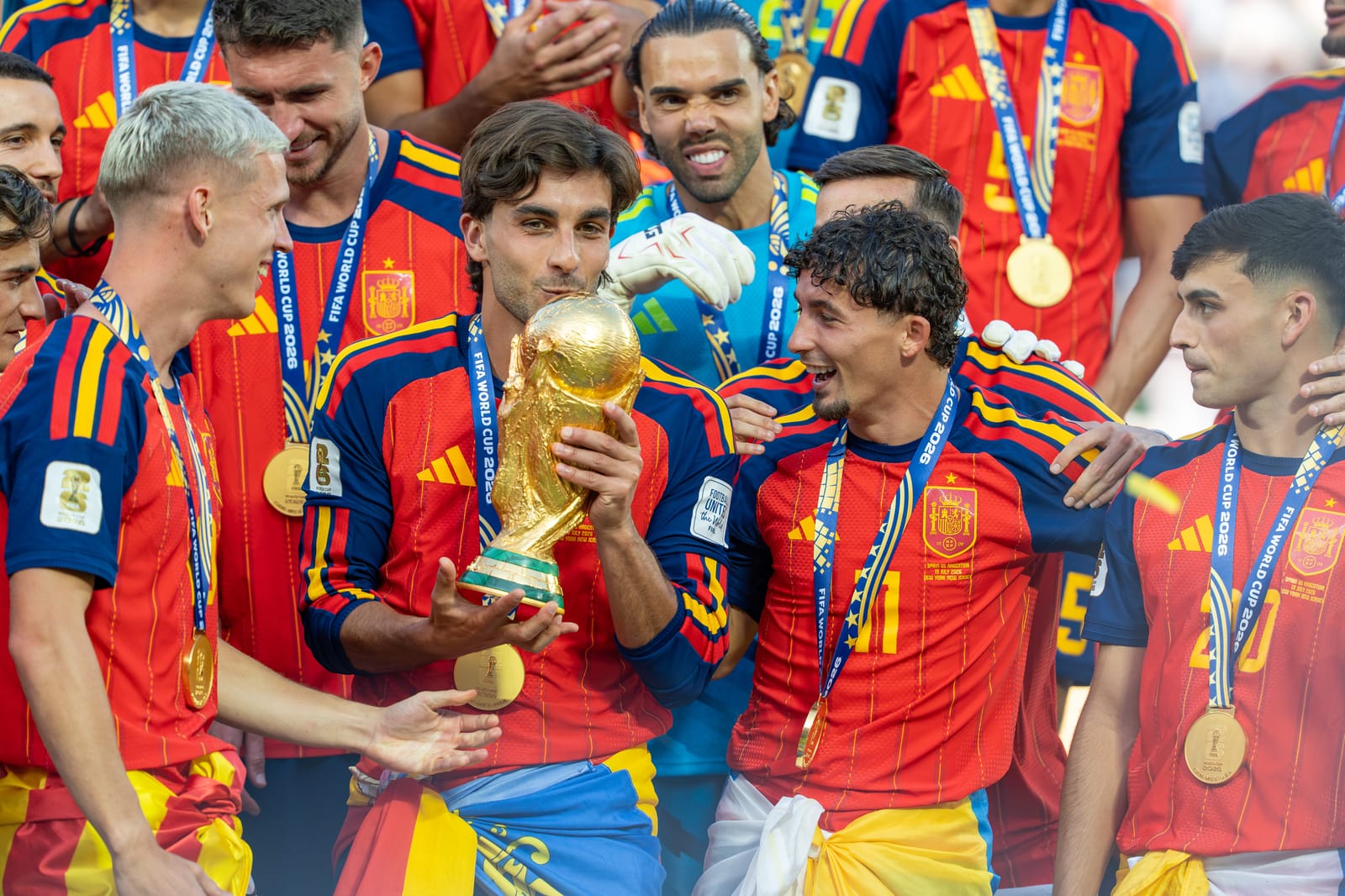 Jugadores de la selección española con camiseta roja y medallas de oro celebran sobre el césped; uno de ellos besa la Copa del Mundo mientras sus compañeros sonríen alrededor.