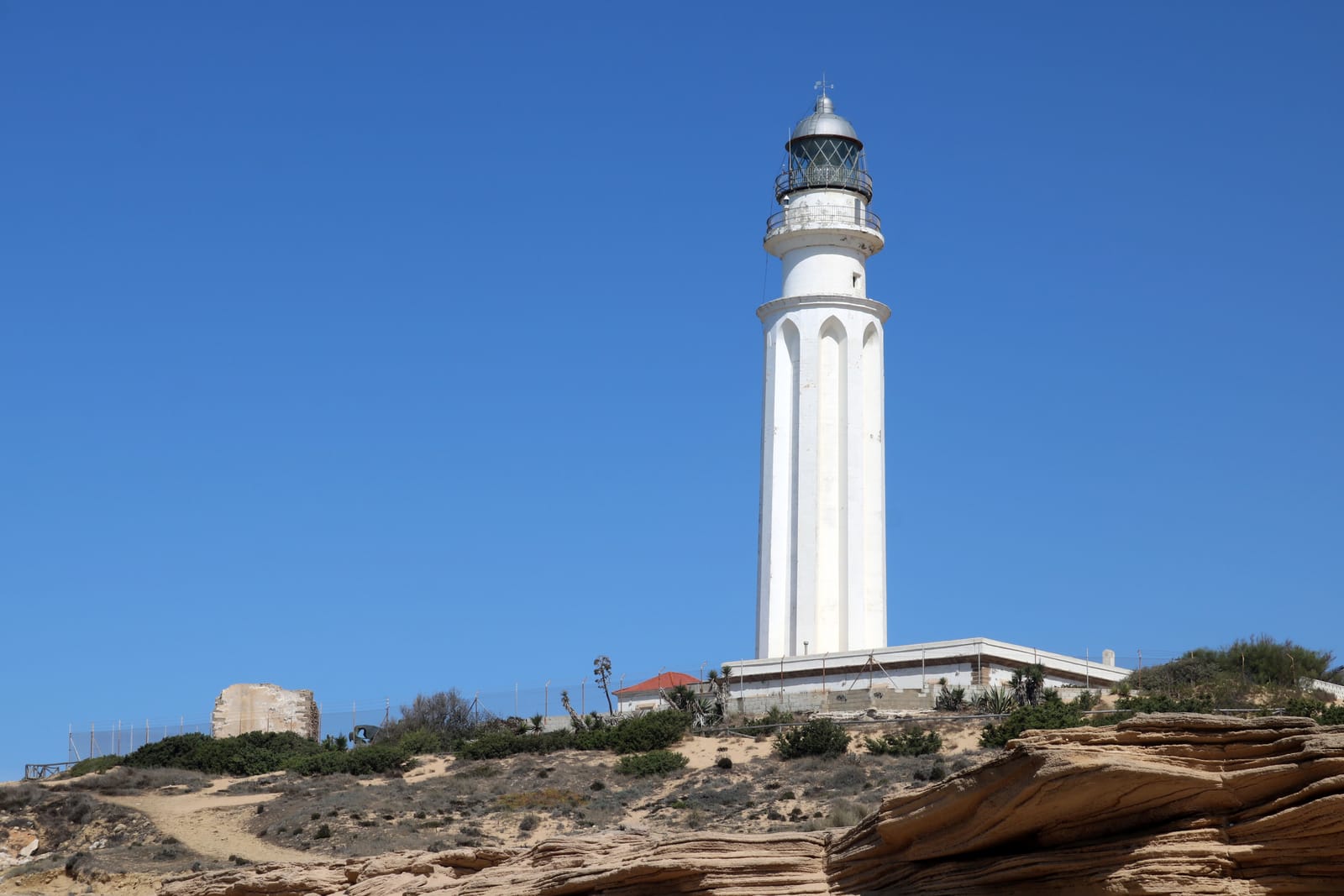 Torre blanca del Faro de Trafalgar sobre un promontorio de arenisca, con un cielo azul despejado y, a la izquierda, los restos de una antigua torre de piedra.