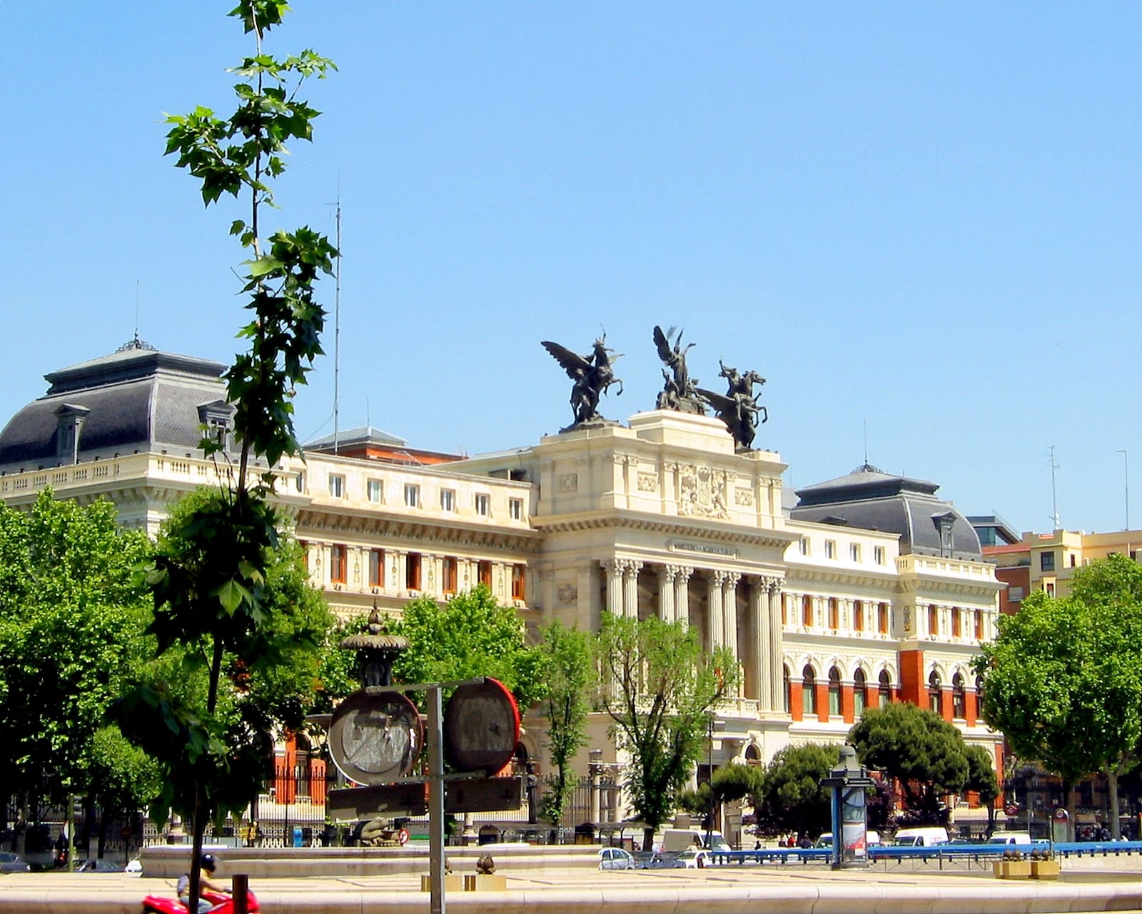 Fachada del Ministerio de Agricultura en Madrid, con columnas, esculturas ecuestres aladas en lo alto y árboles en primer plano bajo un cielo azul.