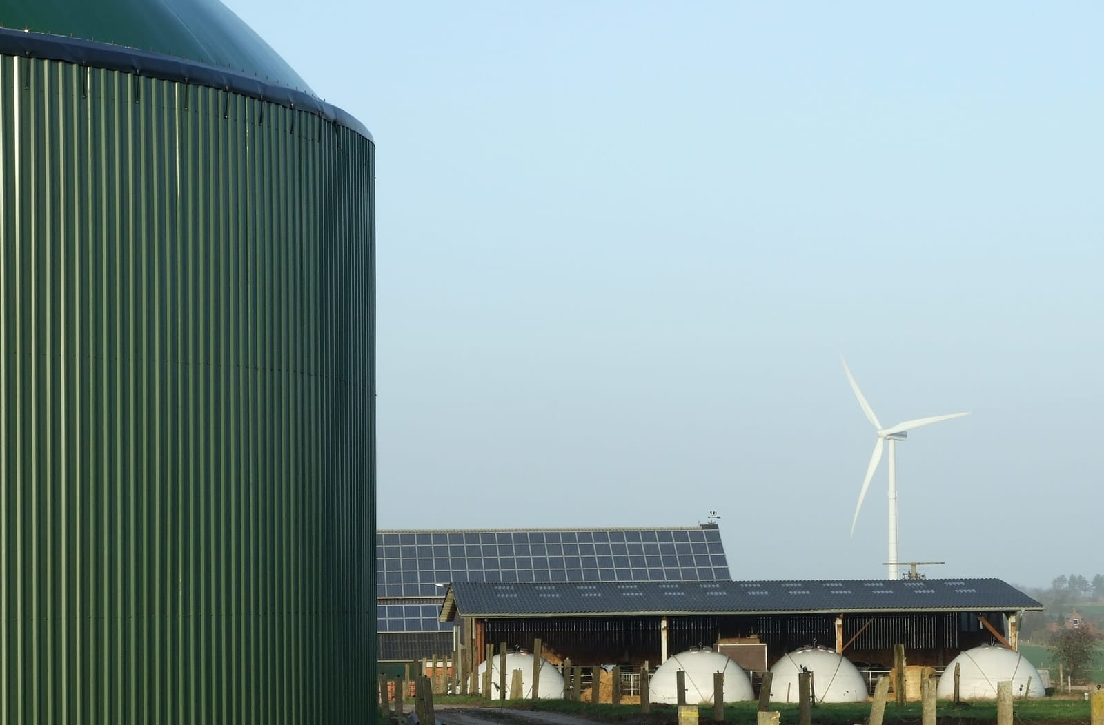 Depósito cilíndrico verde de una planta de biogás en primer plano, con un aerogenerador, una nave con paneles solares y cúpulas blancas de almacenamiento al fondo, en una granja.