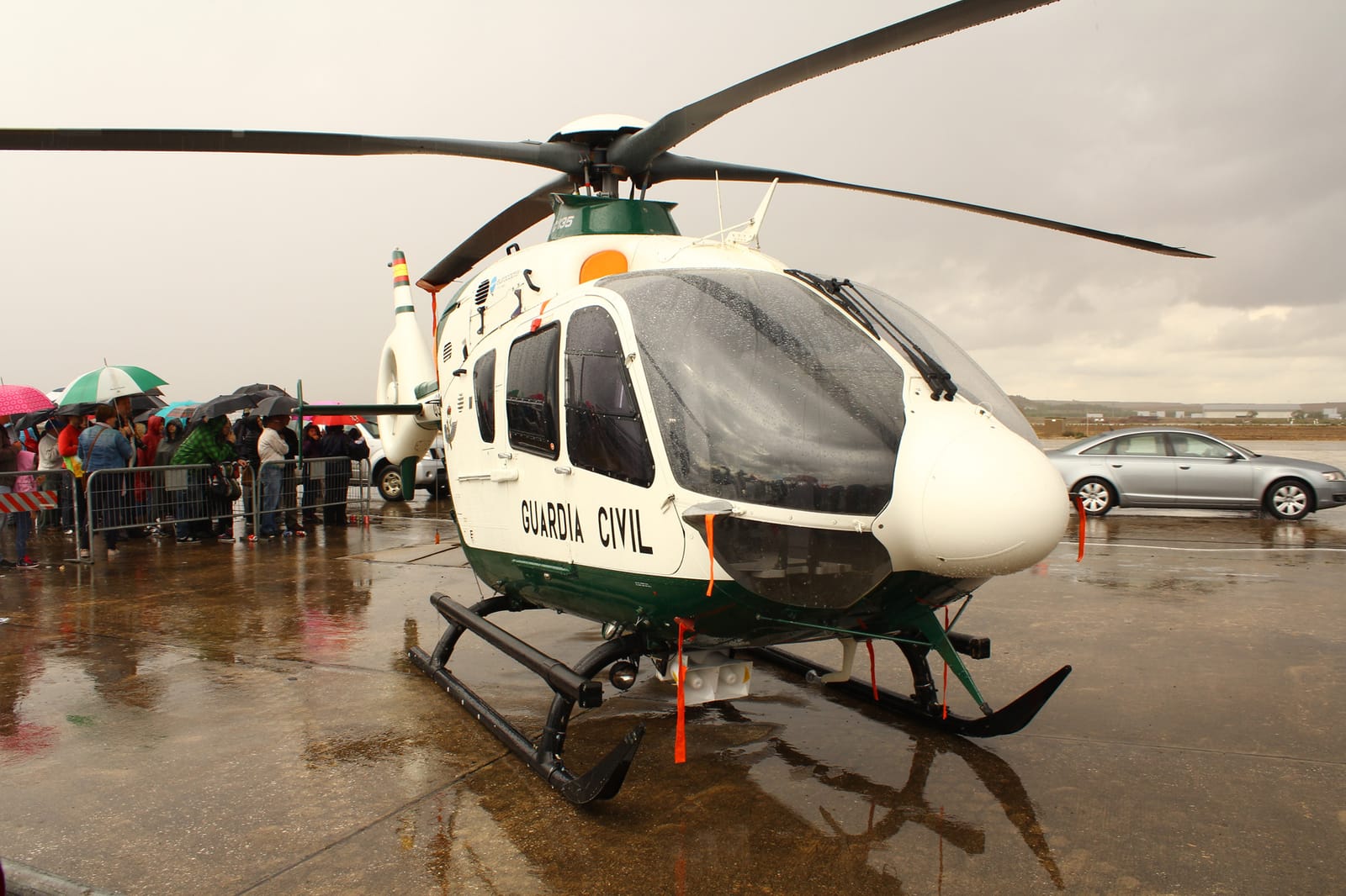Helicóptero Eurocopter EC135 blanco y verde del Servicio Aéreo de la Guardia Civil, posado sobre una pista mojada por la lluvia, con público protegido con paraguas al fondo.