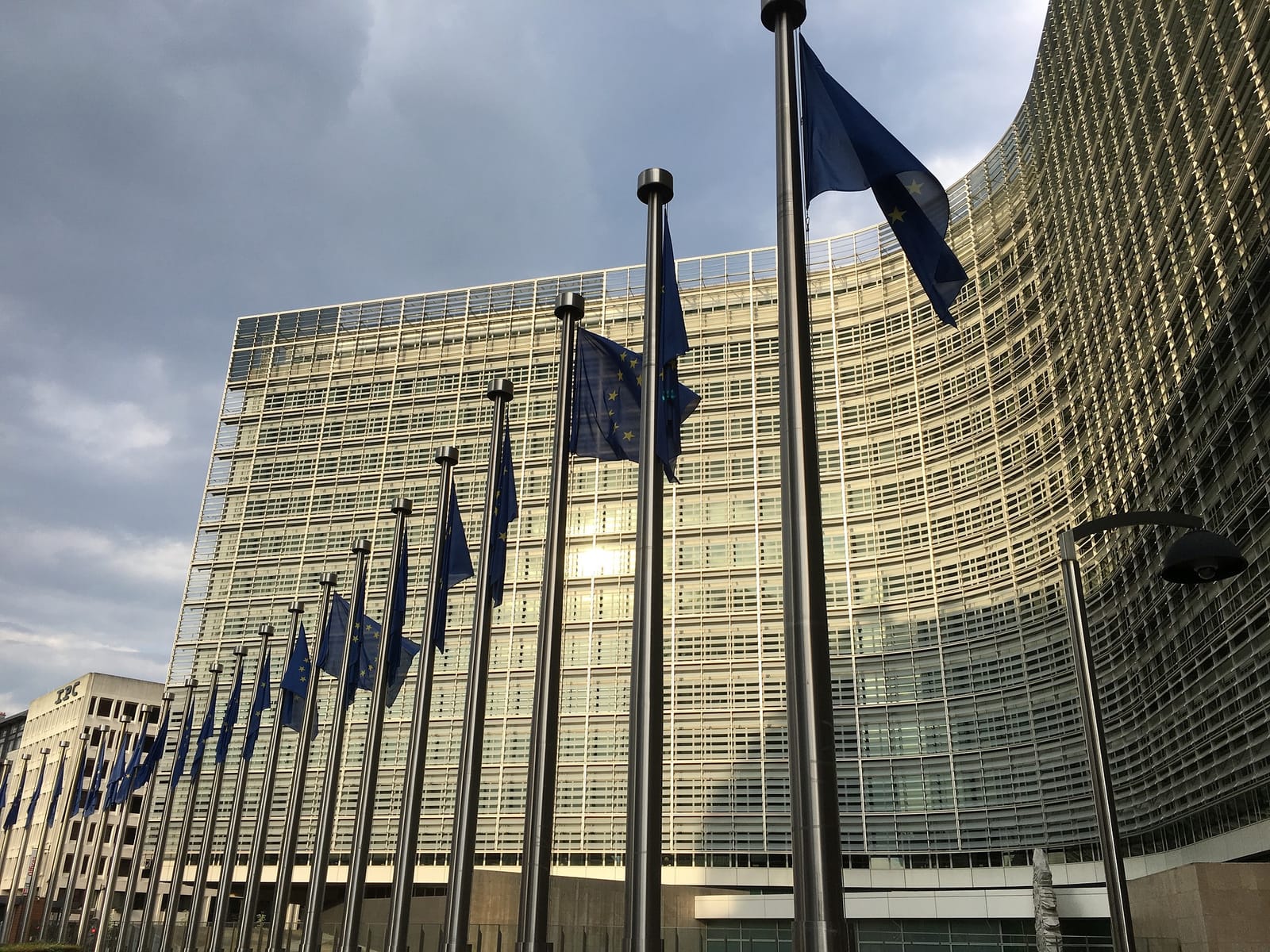 Fachada curva y acristalada del edificio Berlaymont de la Comisión Europea en Bruselas, con una hilera de astas con banderas de la Unión Europea en primer plano y un cielo nublado.