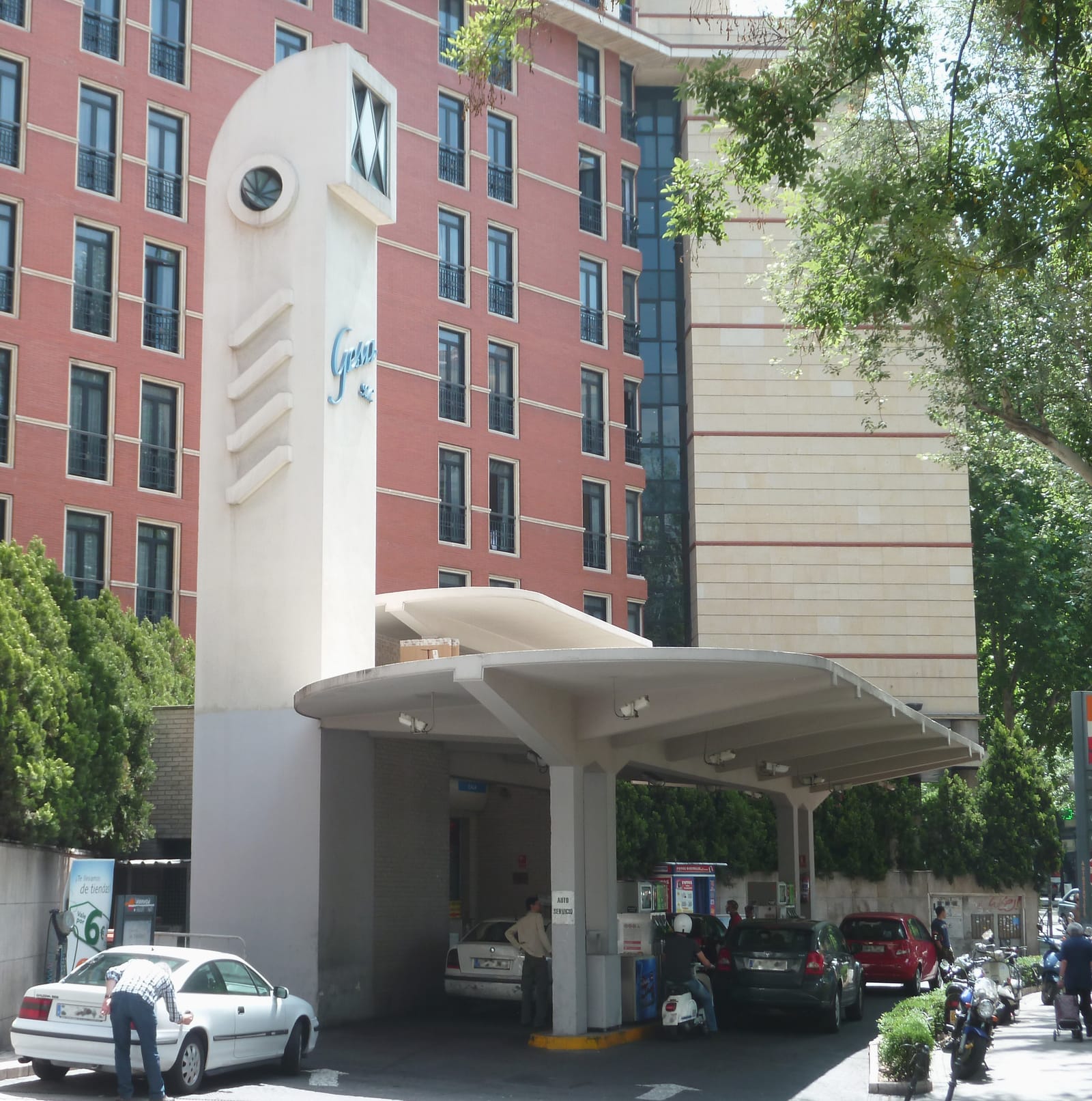 Una gasolinera de Madrid con marquesina blanca y un torreón con el rótulo Gesa, varios coches repostando y bloques de oficinas de ladrillo rojo al fondo.