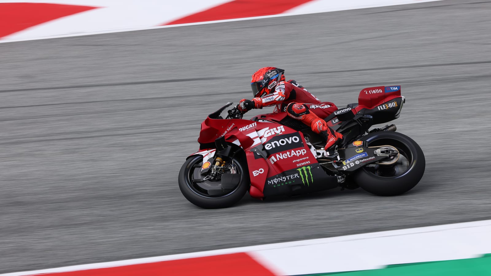 Una moto Ducati roja del equipo oficial, pilotada por un piloto con casco rojo, inclinada en una curva de un circuito con pianos rojiblancos.