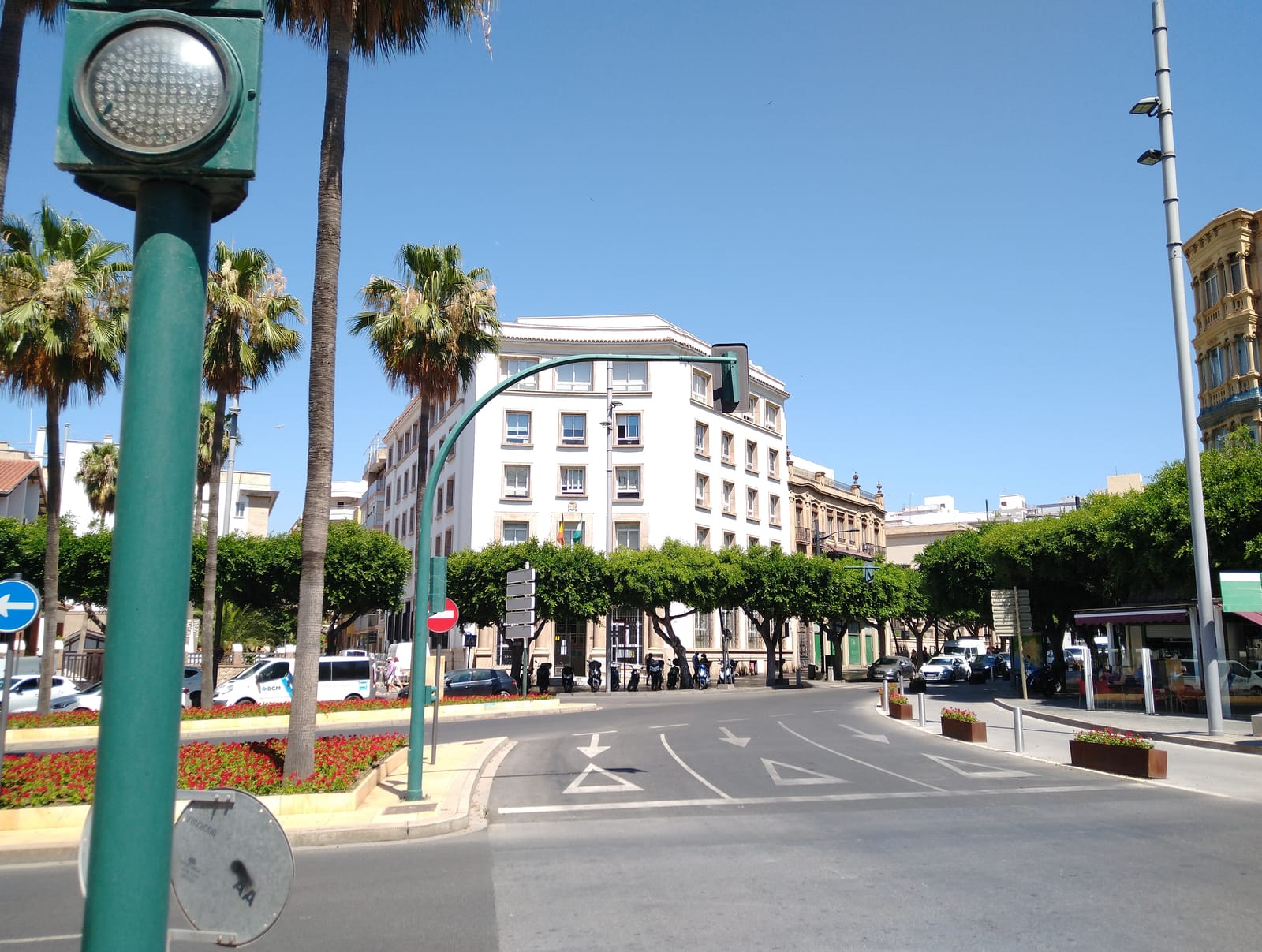 Edificio de oficinas de color claro con fachada de ventanas regulares en una plaza con palmeras, farolas y semáforos, bajo un cielo azul despejado, en el centro de Almería.