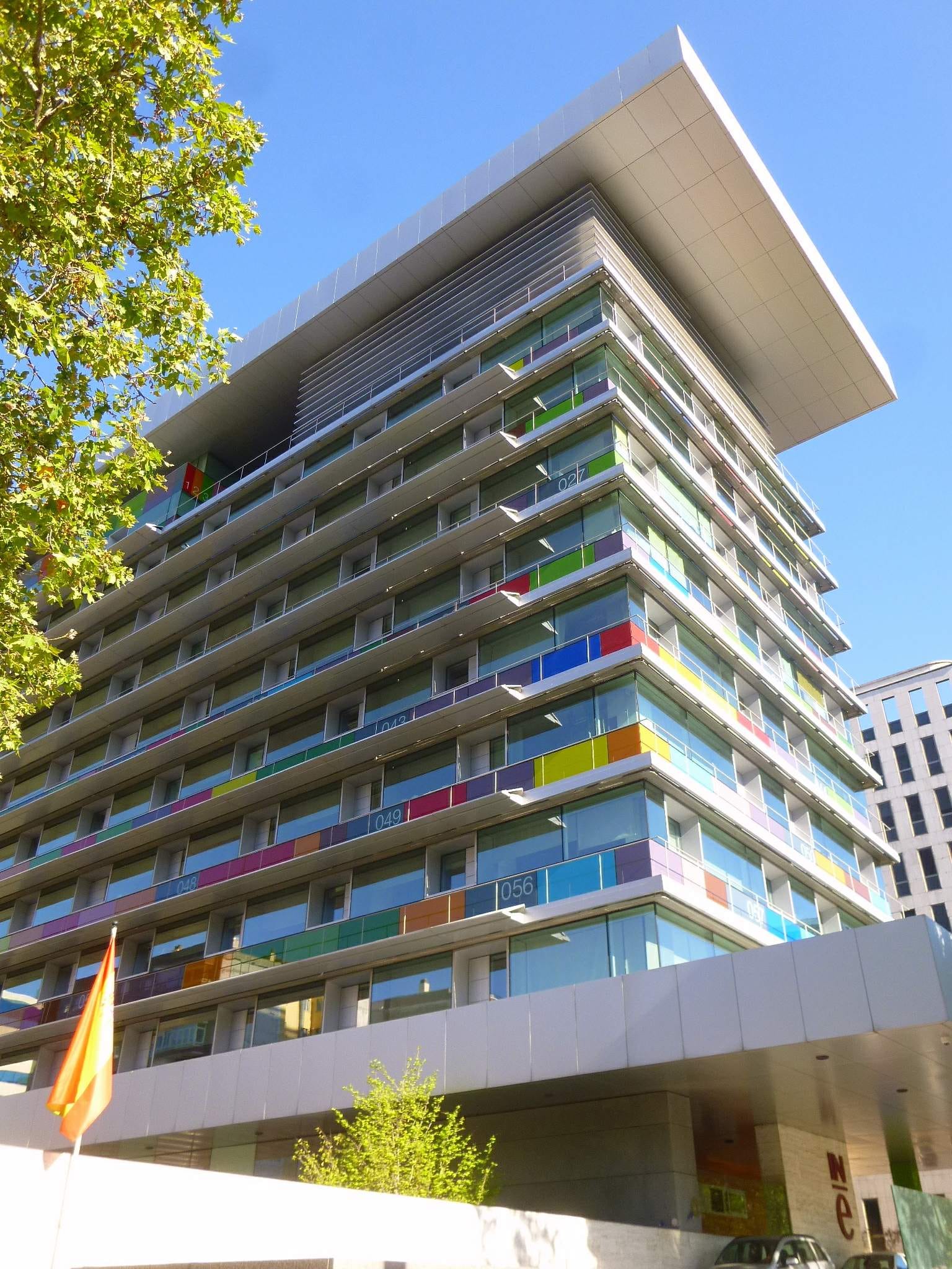 Fachada de un edificio de oficinas de varias plantas con paneles de colores en los balcones, una bandera de España en primer plano y el logotipo del INE en la entrada.