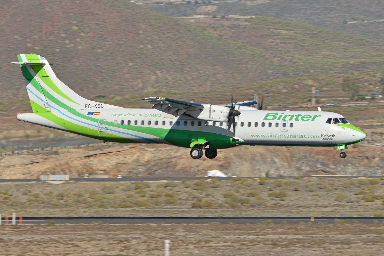 Avión turbohélice ATR 72 blanco y verde de Binter Canarias, matrícula EC-KSG, aterrizando en una pista del aeropuerto de Tenerife Sur con colinas áridas al fondo.
