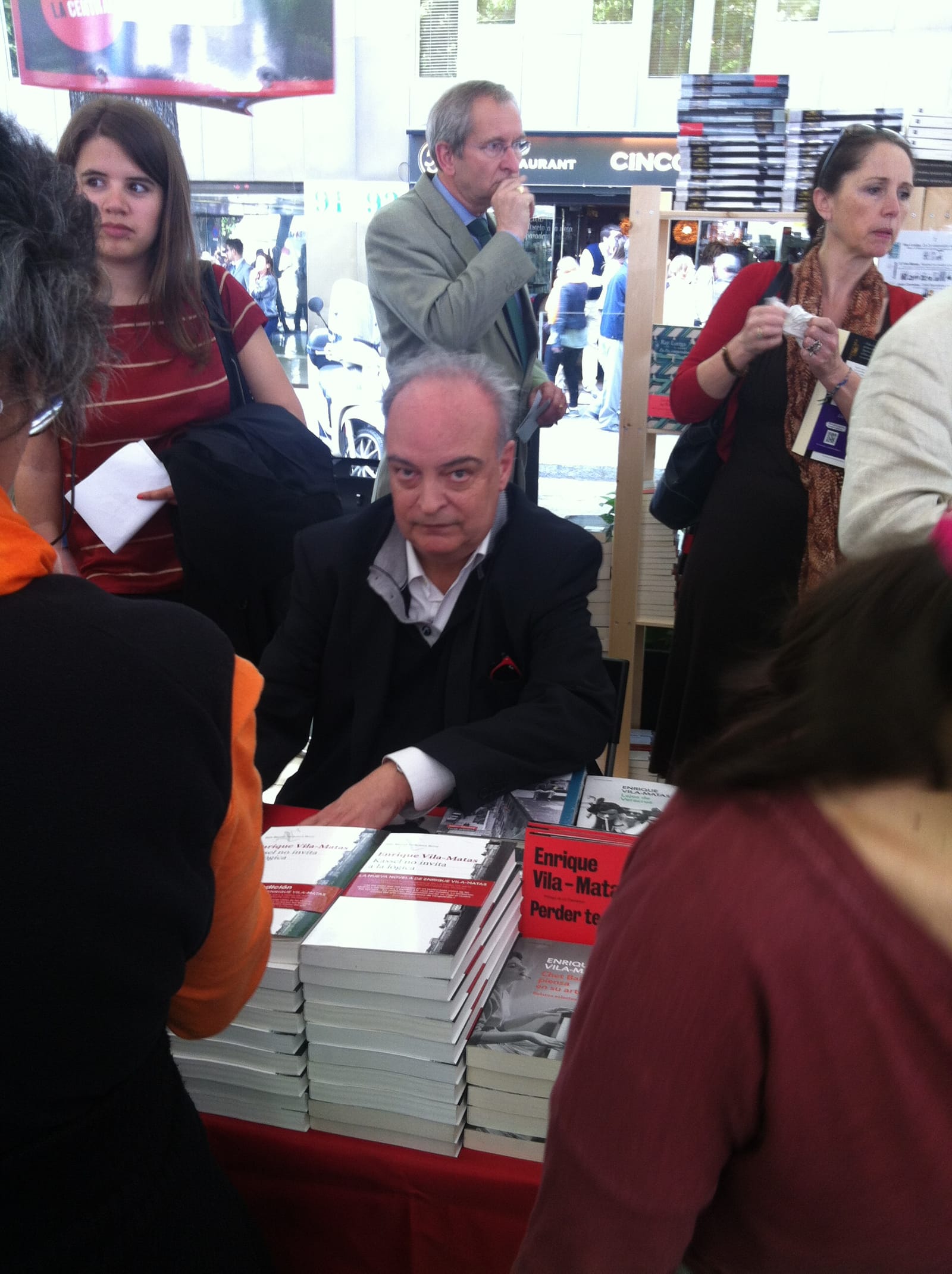Enrique Vila-Matas sentado ante una mesa con pilas de sus libros, rodeado de lectores, durante una firma de ejemplares al aire libre.